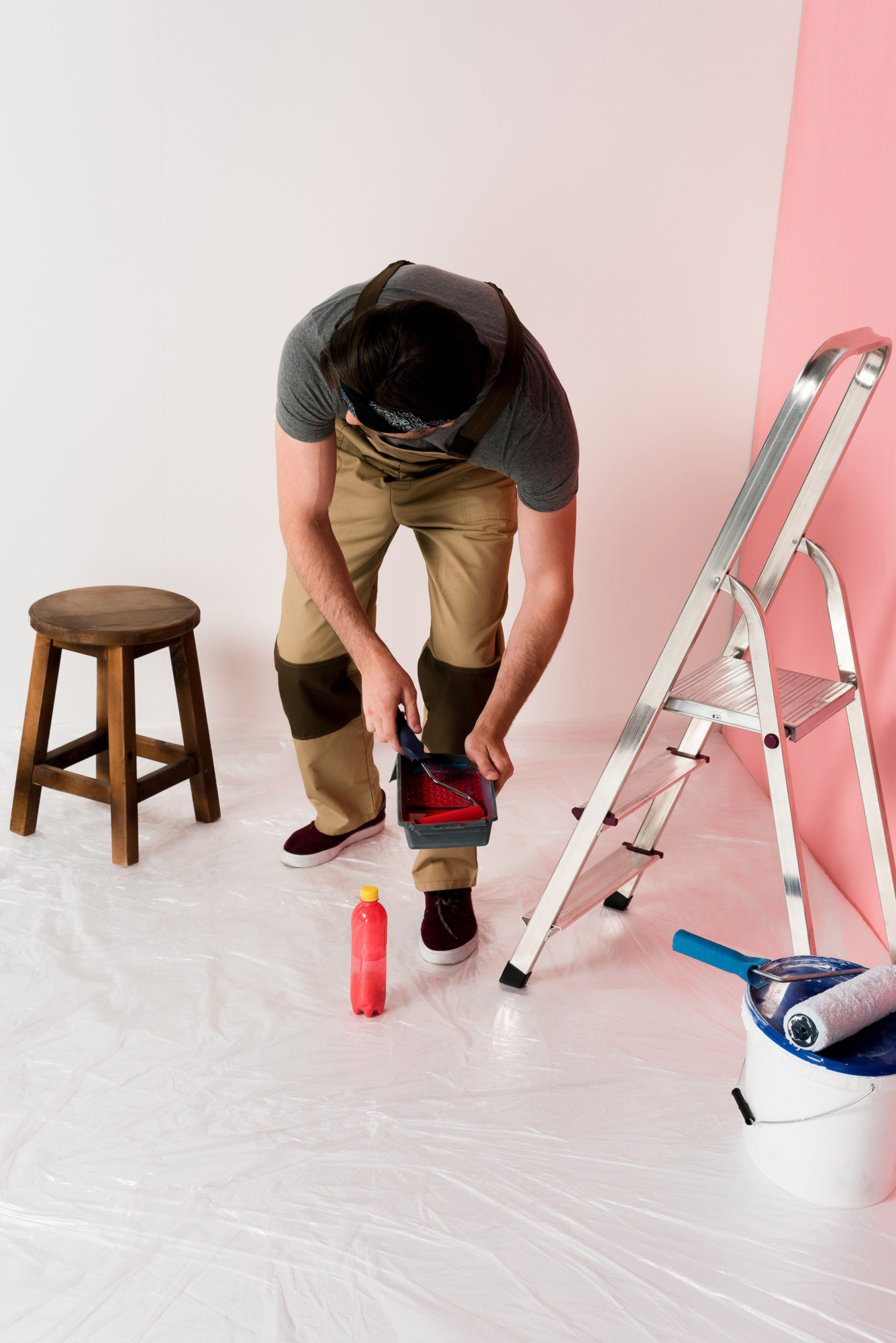 Fotografia de um pintor com o corpo levemente inclinado para baixo, segurando uma bandeja com tinta vermelha com uma mão e um rolo de pintura com a outra mão. Ao seu lado esquerdo há um banquinho de madeira e ao lado direito há uma escada de metal. Em sua frente há uma garrafa de tinta vermelha e uma lata de tinta. Há um plástico protegendo o chão para o início da pintura.