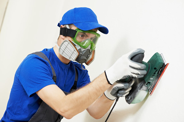 Fotografia de um pintor utilizando máscara, luvas e óculos de proteção enquanto utiliza uma lixadeira