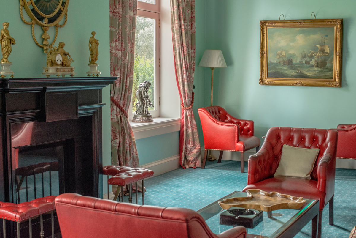 Fotografia de uma sala de estar com as paredes verde-claro. Há diversas poltronas vermelhas, um quadro com moldura dourada, uma lareira em madeira, um janela com cortinas nos tons de branco e vermelho e um carpete azul.