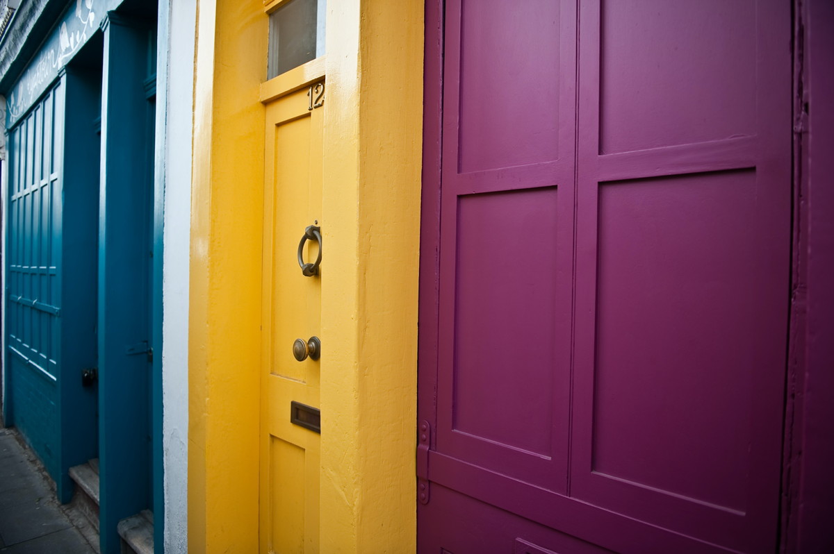 Fotografia de três portas, uma ao lado da outra, nas cores azul, amarelo e roxo.