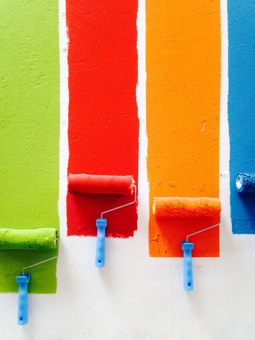 imagem de quatro rolos de pintura apoiados sobre o chão. Cada um está sujo de uma cor diferente (verde, vermelho, laranja e azul) e sobre uma faixa da respectiva cor.