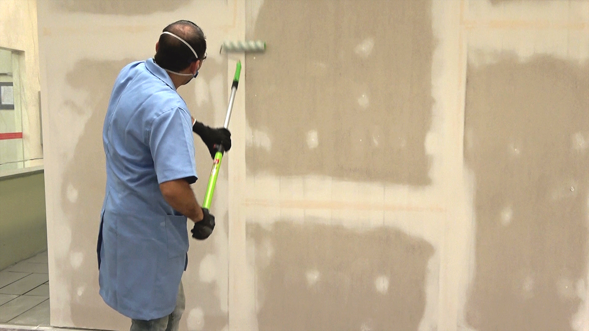 Fotografia de um homem pintando uma parede. Ele utiliza luvas, máscara de proteção e um rolo de pintura acoplado a um cabo extensor.