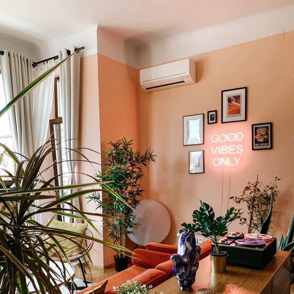 Fotografia de uma sala de estar. Há um sofá vermelho, um aparador em madeira, objetos de decoração (sobre ele e pendurados na parede), um aparelho de ar-condicionado e plantas. As paredes são laranja.