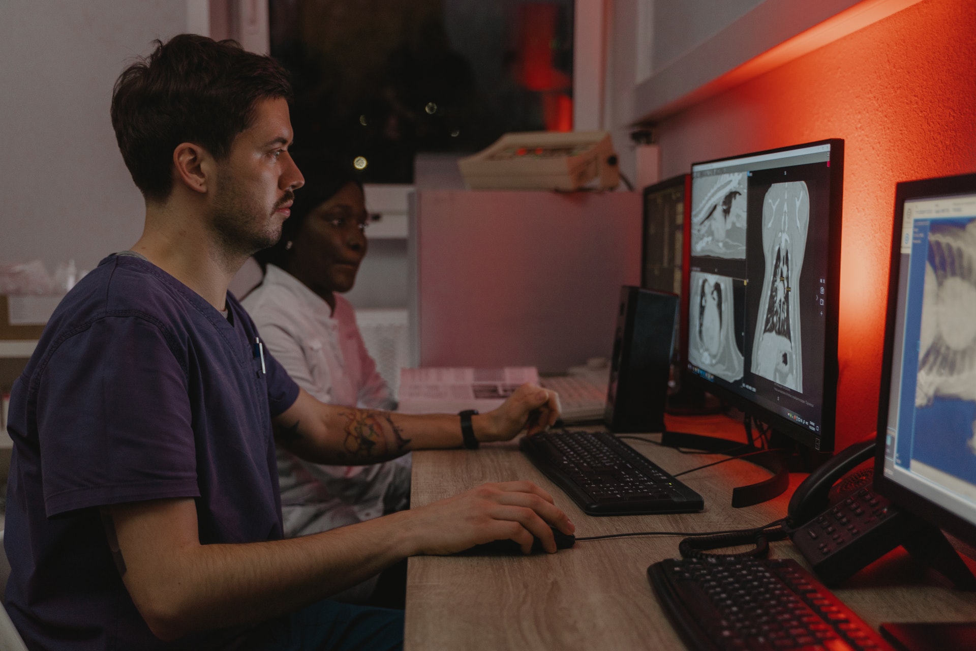 Foto de duas pessoas utilizando computadores em um ambiente como escritório. Em primeiro plano, aparece a imagem de um homem segurando o mouse e olhando para tela do computador; em segundo plano, há uma mulher também olhando para a tela do computador.