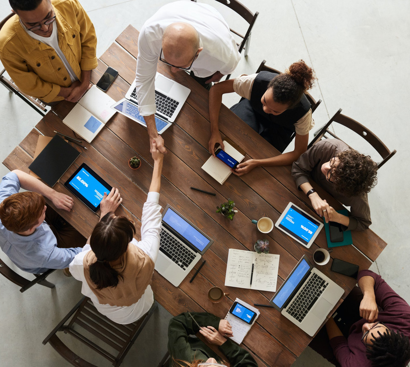 Foto de uma mesa retangular com seis pessoas sentadas em seu entorno, tirada de cima. Sobre a mesa, há vários aparelhos tecnológicos, como: smartphones, tablets e notebooks. Há também cadernos, canetas e duas xicaras de café. Uma das pessoas sentadas, um homem branco, vestindo camisa branca, aperta a mão de uma mulher branca, vestida de camisa branca e xale bege. As demais pessoas são jovens: dois brancos e dois jovens negros.