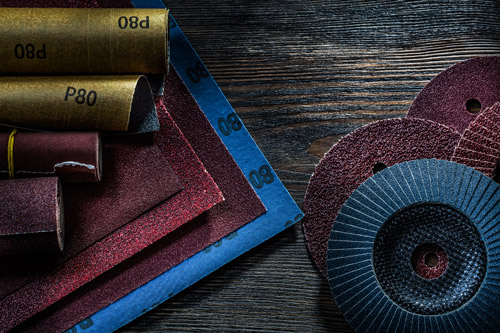 Fotografia de diversas lixas com diferentes gramaturas, formatos e cores dispostas sobre uma mesa. Algumas estão enroladas e presas com um elástico e outras estão sobrepostas umas às outras.