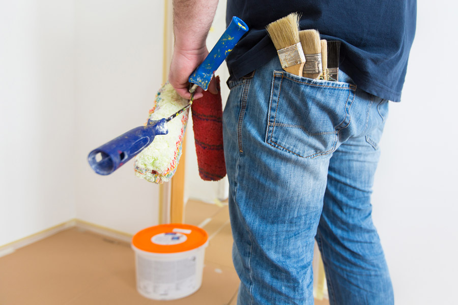 Fotografia de um homem de costas segurando dois rolos de pintura. É possível ver um pote plástico tampado no chão e diversos pincéis no bolso traseiro da calça dele. 