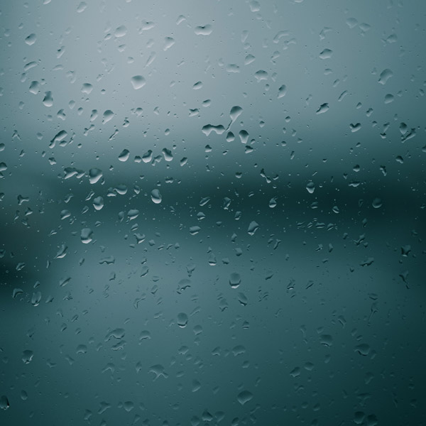 Fotografia de gotas de água em uma superfície de vidro. O vidro está embaçado e é possível identificar apenas que atrás dele há uma paisagem em tons de cinza.