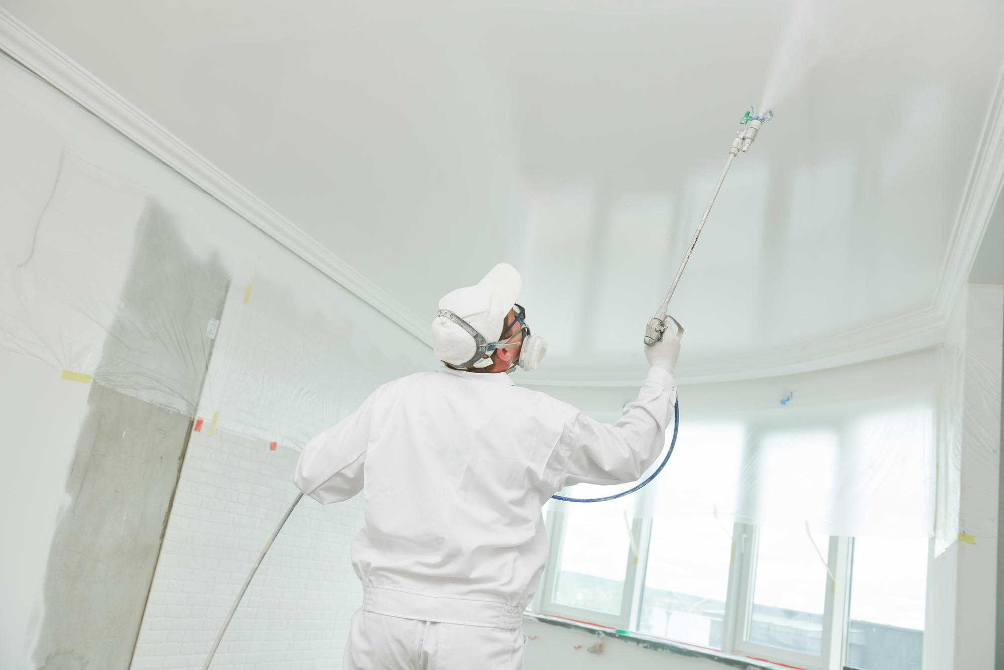 Fotografia de um homem pintando o teto de um imóvel de branco com o auxílio de um equipamento pulverizador. Ele usa EPIs (máscara, óculos e luvas).