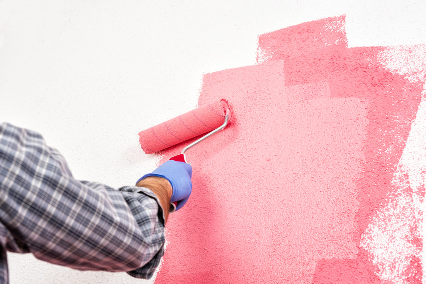 Fotografia de uma pessoa pintando uma parede branca de rosa. Ela utiliza luvas e um rolo de lã.