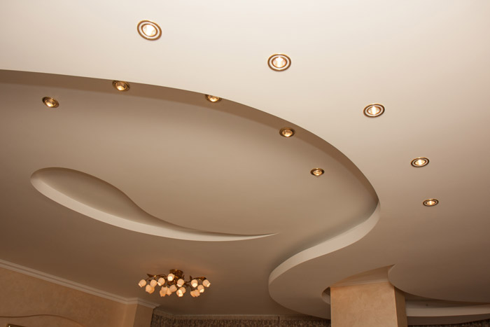 Fotografia de um teto com aplicação de drywall finalizado. No drywall há desníveis e recortes curvilíneos e pontos de ilumação.